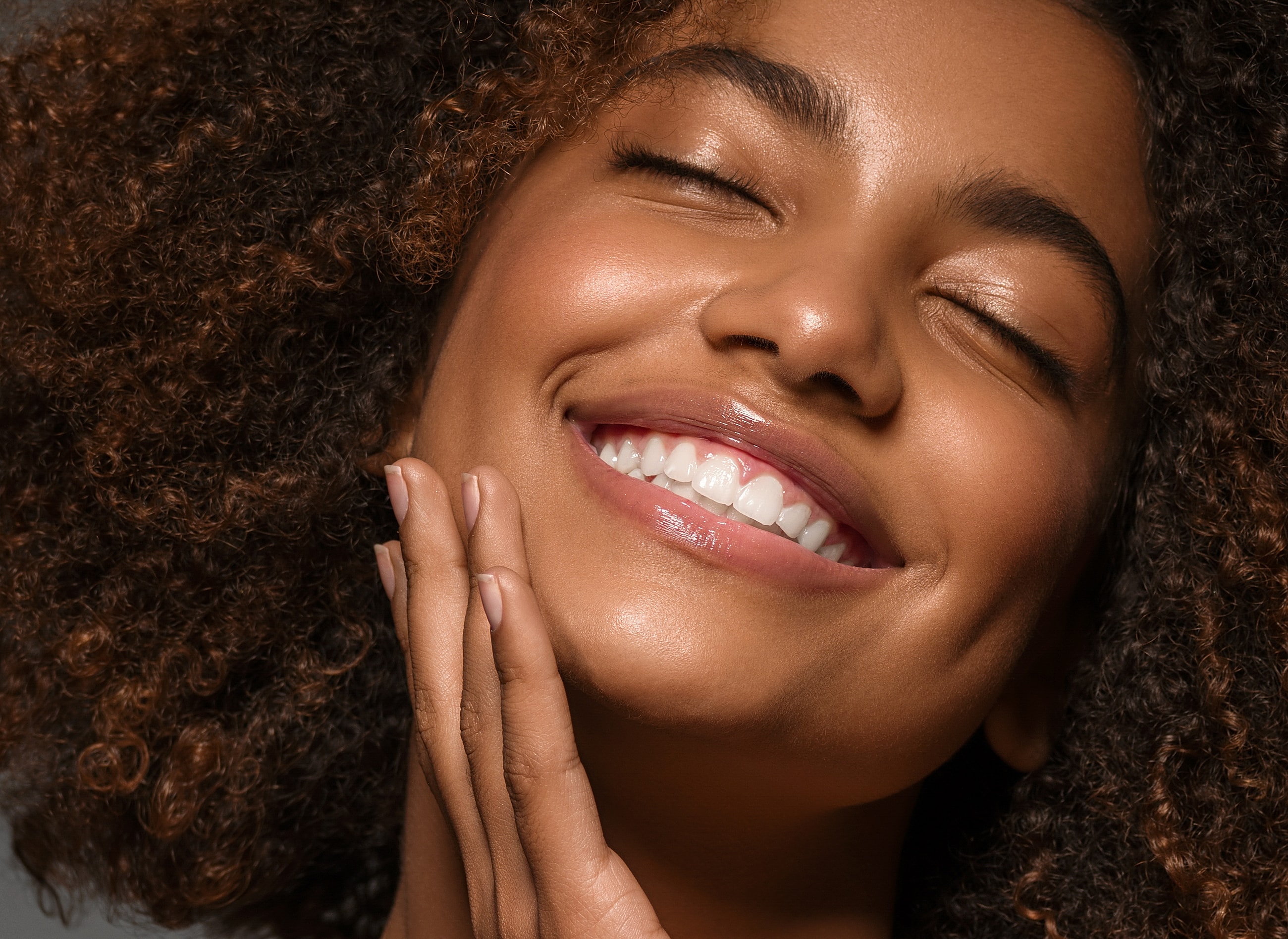 Smiling moxi laser patient model with curly hair and glowing skin touching her face